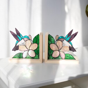 Pu&ograve; includere: Un paio di fermalibri in legno con disegni di colibr&igrave; e fiori in vetro colorato. I colibr&igrave; hanno dettagli turchese, viola e bianchi, mentre i fiori sono color crema con foglie verdi. I fermalibri sono appoggiati su una superficie bianca.