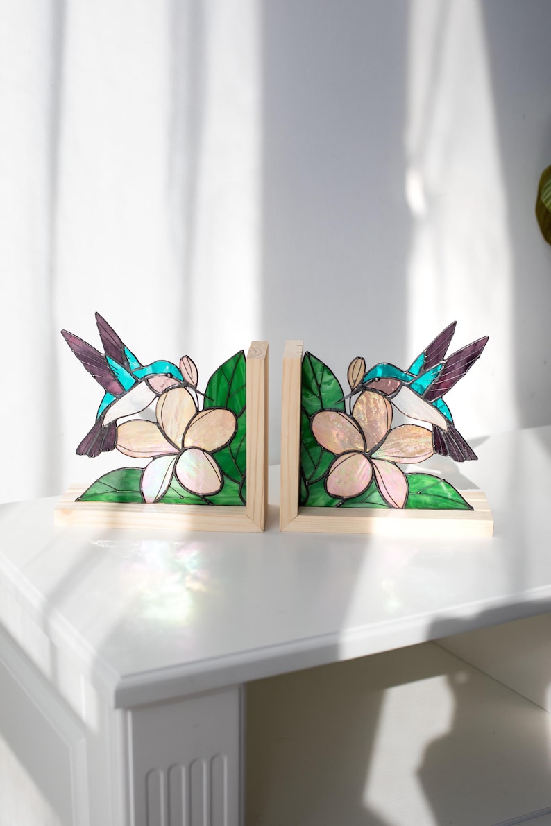 Pu&ograve; includere: Un paio di fermalibri in legno con disegni di colibr&igrave; e fiori in vetro colorato. I colibr&igrave; hanno dettagli turchese, viola e bianchi, mentre i fiori sono color crema con foglie verdi. I fermalibri sono appoggiati su una superficie bianca.