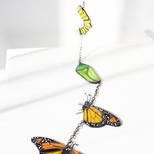 May include: A decorative hanging mobile featuring a monarch butterfly life cycle. The mobile includes a yellow egg, a green chrysalis, and two orange and black butterflies, all connected by a chain.