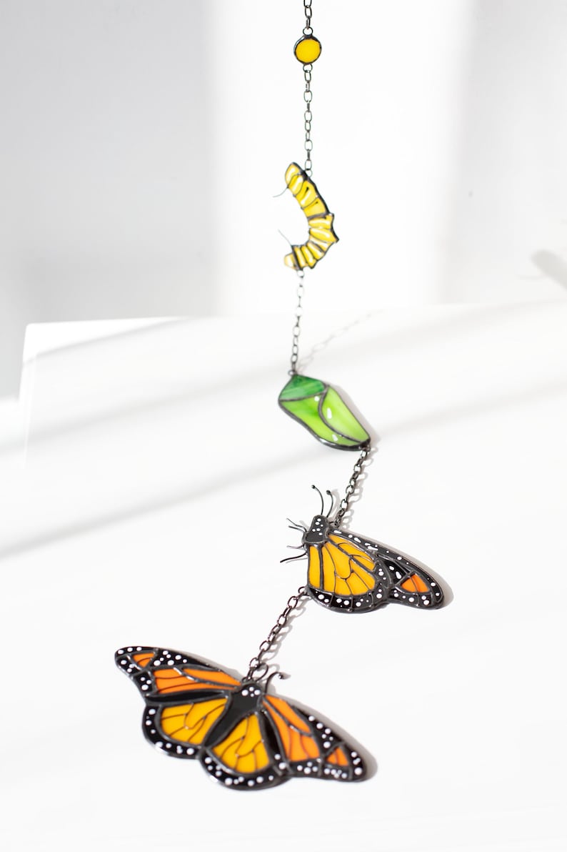 May include: A decorative hanging mobile featuring a monarch butterfly life cycle. The mobile includes a yellow egg, a green chrysalis, and two orange and black butterflies, all connected by a chain.