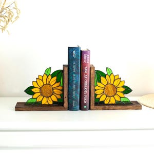 May include: Wooden bookends featuring a stained glass sunflower design. The sunflowers have yellow petals and green leaves. Two books stand between the bookends. The bookends are on a white surface.