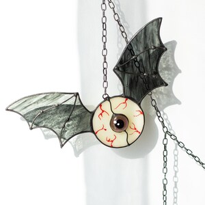 May include: A stained glass suncatcher shaped like a bat with a central eyeball design. The bat wings are made of translucent grey glass with dark veining, while the central eye features a white sclera with red veins and a dark brown pupil.