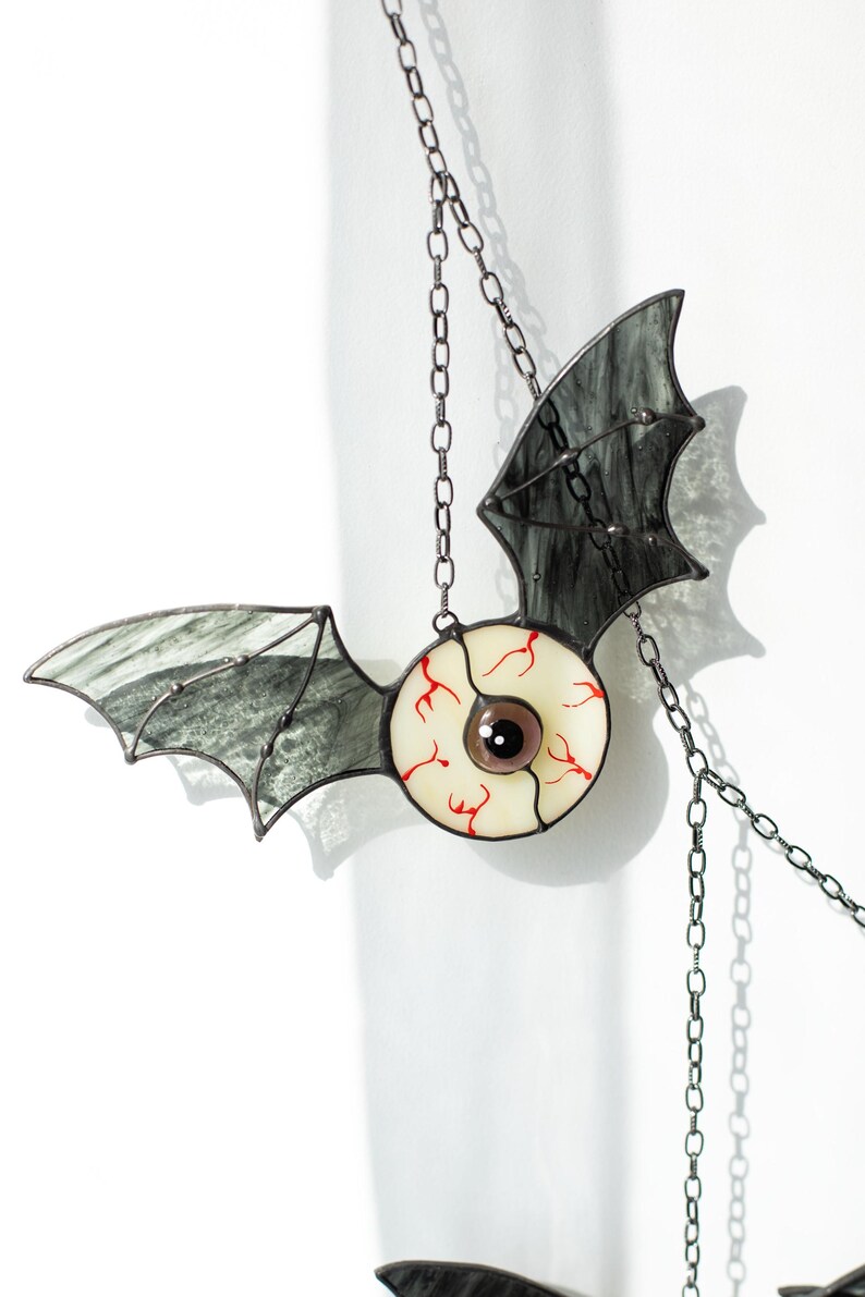 May include: A stained glass suncatcher shaped like a bat with a central eyeball design. The bat wings are made of translucent grey glass with dark veining, while the central eye features a white sclera with red veins and a dark brown pupil.