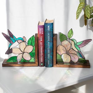 Pu&ograve; includere: Reggilibri in vetro colorato con un design di colibr&igrave; e fiori. I reggilibri sono realizzati in legno e vetro, con un colibr&igrave; nei toni del blu, verde e viola. Il fiore &egrave; di colore rosa chiaro. Due libri sono posizionati tra i reggilibri.
