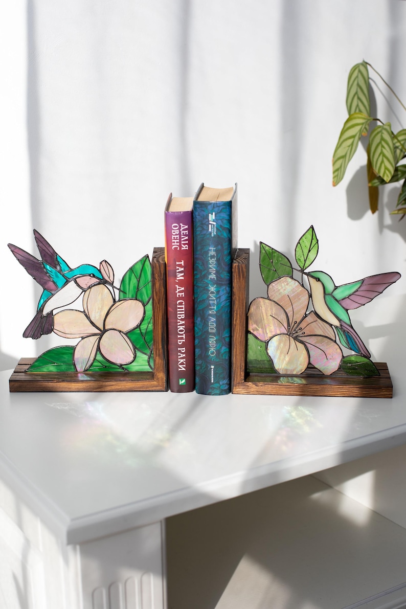 Pu&ograve; includere: Reggilibri in vetro colorato con un design di colibr&igrave; e fiori. I reggilibri sono realizzati in legno e vetro, con un colibr&igrave; nei toni del blu, verde e viola. Il fiore &egrave; di colore rosa chiaro. Due libri sono posizionati tra i reggilibri.