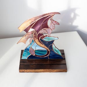 Op de afbeelding: Een gebrandschilderde drakenbeeldhouwwerk op een donkere houten basis. De draak heeft paarse vleugels en een lichtroze lichaam, geplaatst op een blauwe en groene glazen formatie. Het is een decoratief item.