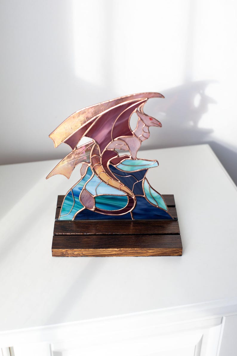 Op de afbeelding: Een gebrandschilderde drakenbeeldhouwwerk op een donkere houten basis. De draak heeft paarse vleugels en een lichtroze lichaam, geplaatst op een blauwe en groene glazen formatie. Het is een decoratief item.