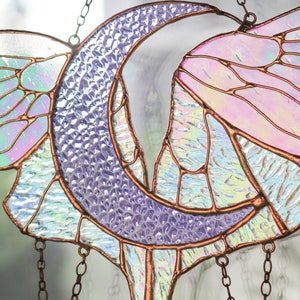 May include: A stained glass suncatcher featuring a crescent moon and a stylized bird design. The glass is iridescent and has a purple, pink, and white color scheme. The suncatcher is hung from a copper chain.