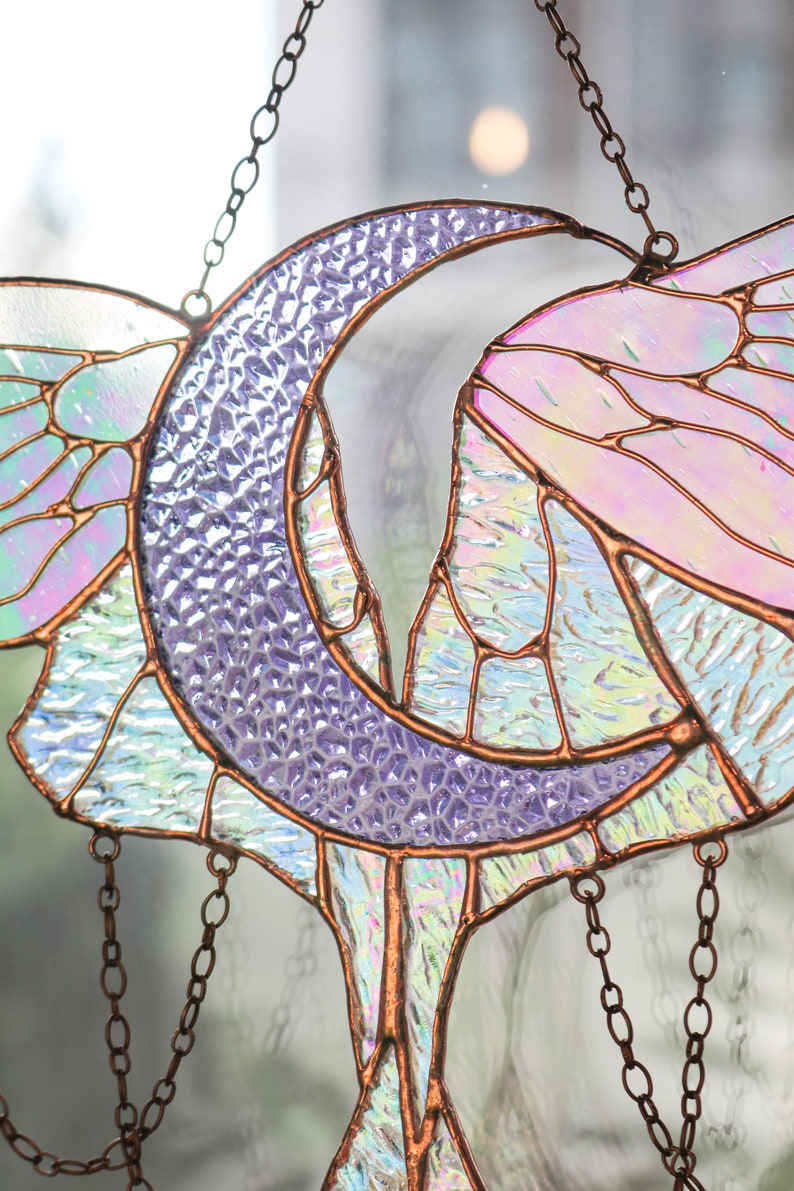 May include: A stained glass suncatcher featuring a crescent moon and a stylized bird design. The glass is iridescent and has a purple, pink, and white color scheme. The suncatcher is hung from a copper chain.