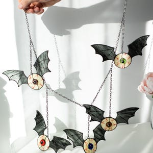 May include: A hanging mobile featuring four stained glass bat ornaments, each with a central eyeball design. The bats have dark grey wings and irises in green, brown, red, and blue, all connected by thin metal chains for display.