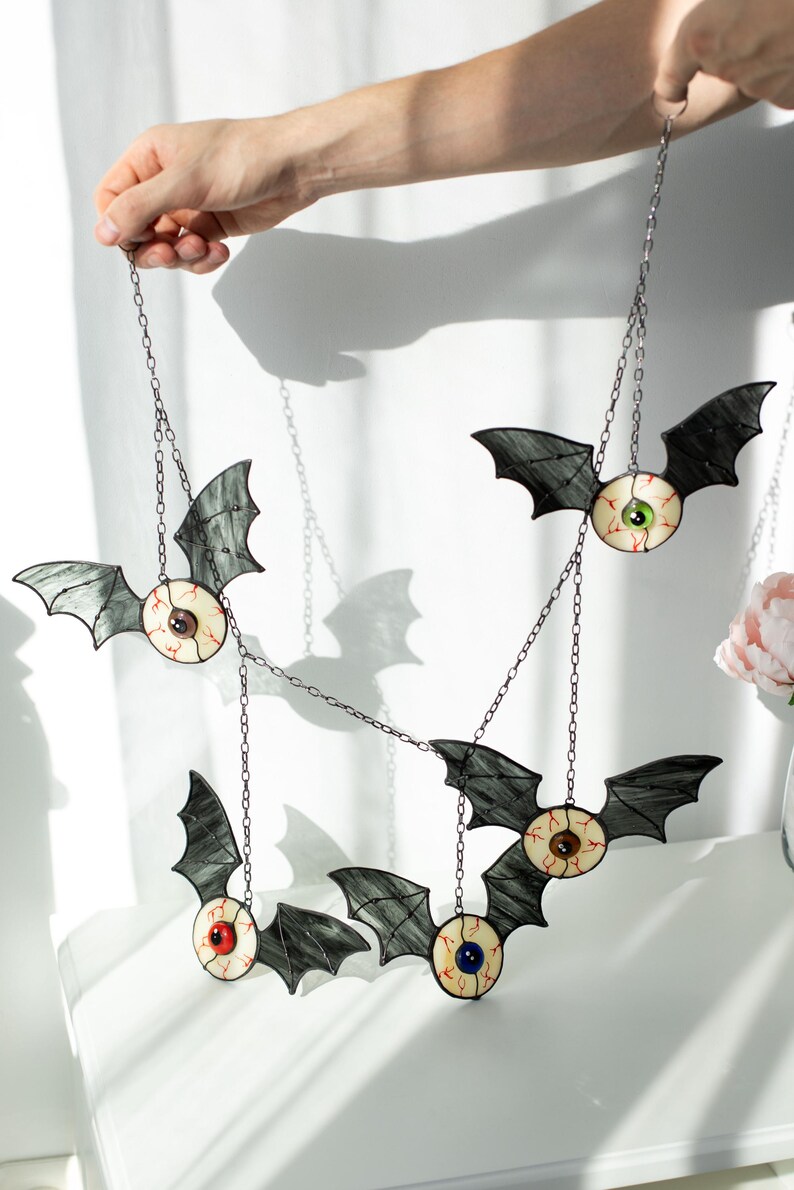 May include: A hanging mobile featuring four stained glass bat ornaments, each with a central eyeball design. The bats have dark grey wings and irises in green, brown, red, and blue, all connected by thin metal chains for display.