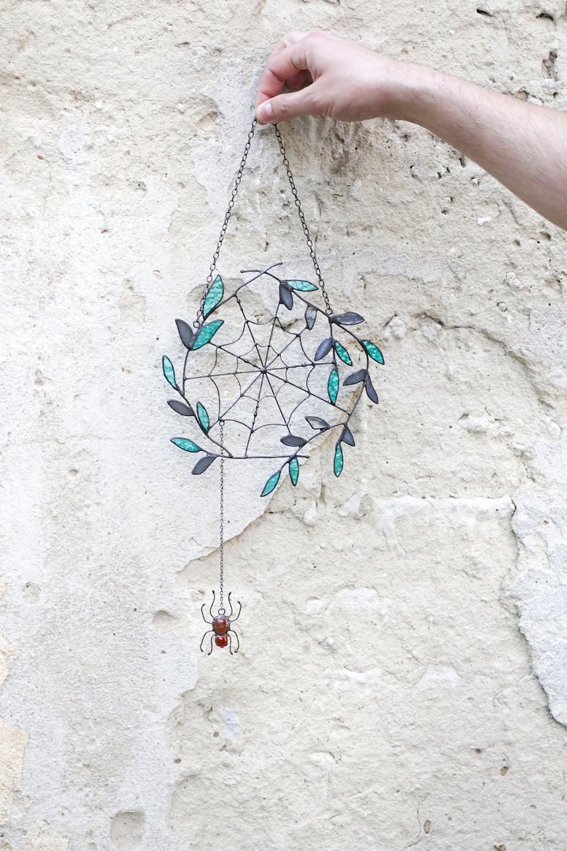 May include: A black metal spider web with teal glass leaves hanging from a chain with a small metal spider dangling below.