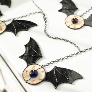 May include: A decorative garland featuring multiple stained glass bat ornaments connected by a dark metal chain. Each bat has black wings and a central circular body resembling an eyeball with a white sclera, red veins, and a dark blue pupil.