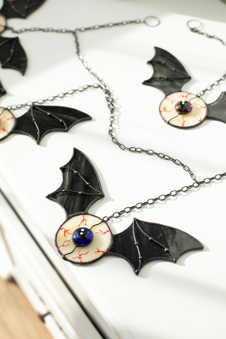 May include: A decorative garland featuring multiple stained glass bat ornaments connected by a dark metal chain. Each bat has black wings and a central circular body resembling an eyeball with a white sclera, red veins, and a dark blue pupil.