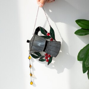 Puede incluir: Un atrapasue&ntilde;os de vidrieras con forma de cafetera moka, que presenta paneles de vidrio gris, hojas de color verde oscuro y peque&ntilde;as bayas de vidrio rojo. Una cadena colgante con cuentas de vidrio de color &aacute;mbar cuelga del pico.