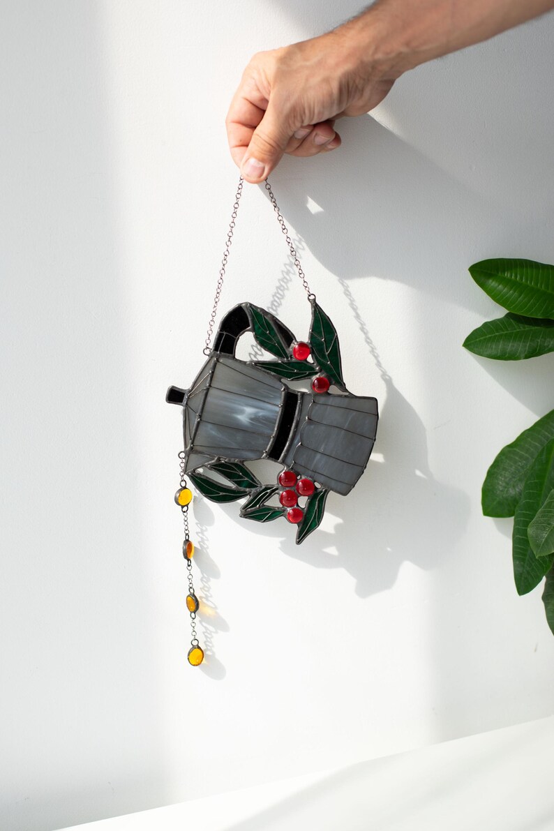 Puede incluir: Un atrapasue&ntilde;os de vidrieras con forma de cafetera moka, que presenta paneles de vidrio gris, hojas de color verde oscuro y peque&ntilde;as bayas de vidrio rojo. Una cadena colgante con cuentas de vidrio de color &aacute;mbar cuelga del pico.