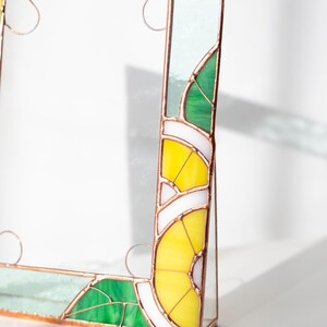 May include: Stained glass picture frame with a botanical design. The frame features green, yellow, white, and clear glass pieces, with copper-colored metal accents. The design includes leaf and lemon slice motifs.