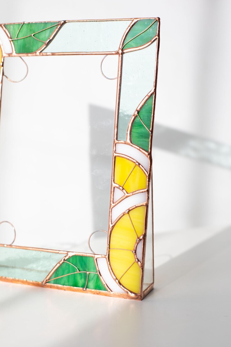 May include: Stained glass picture frame with a botanical design. The frame features green, yellow, white, and clear glass pieces, with copper-colored metal accents. The design includes leaf and lemon slice motifs.