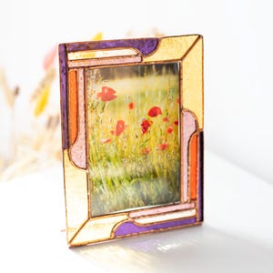 Op de afbeelding: Een glas-in-lood fotolijst met een landschapsfoto van rode klaprozen in een veld. De lijst heeft een goud, paars, oranje en roze kleurenschema. De lijst staat rechtop op een wit oppervlak.