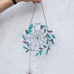 May include: A black metal spider web with teal leaves hanging from a chain with a small spider dangling below. The spider has a red and orange body.