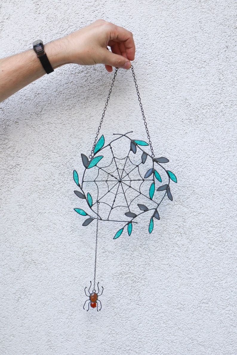 May include: A black metal spider web with teal leaves hanging from a chain with a small spider dangling below. The spider has a red and orange body.
