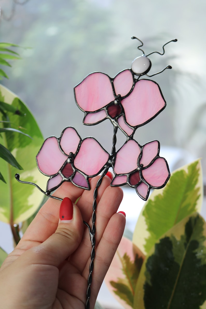 K&ouml;nnte beinhalten: Eine rosafarbene Orchideenbl&uuml;te aus Glasmalerei mit einer wei&szlig;en Perle an der Spitze des Stiels. Die Orchidee besteht aus mehreren Glasscheiben und wird in der Hand einer Person gehalten.