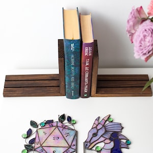 May include: A wooden bookend holds two books with colorful spines. Below, a stained glass D20 die and a dragon head are displayed. The die is multi-colored with numbers, and the dragon is purple and blue.