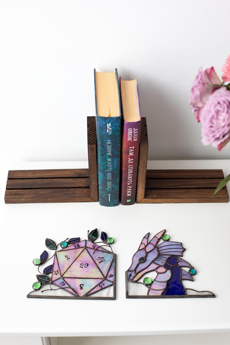 May include: A wooden bookend holds two books with colorful spines. Below, a stained glass D20 die and a dragon head are displayed. The die is multi-colored with numbers, and the dragon is purple and blue.