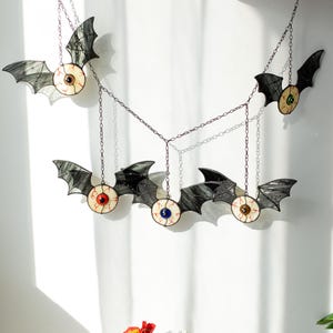 May include: A hanging mobile featuring five stained glass bats, each with a central eyeball design. The bats have dark grey wings and eyes with red, blue, green, brown, and black irises, all connected by thin metal chains for suspension.