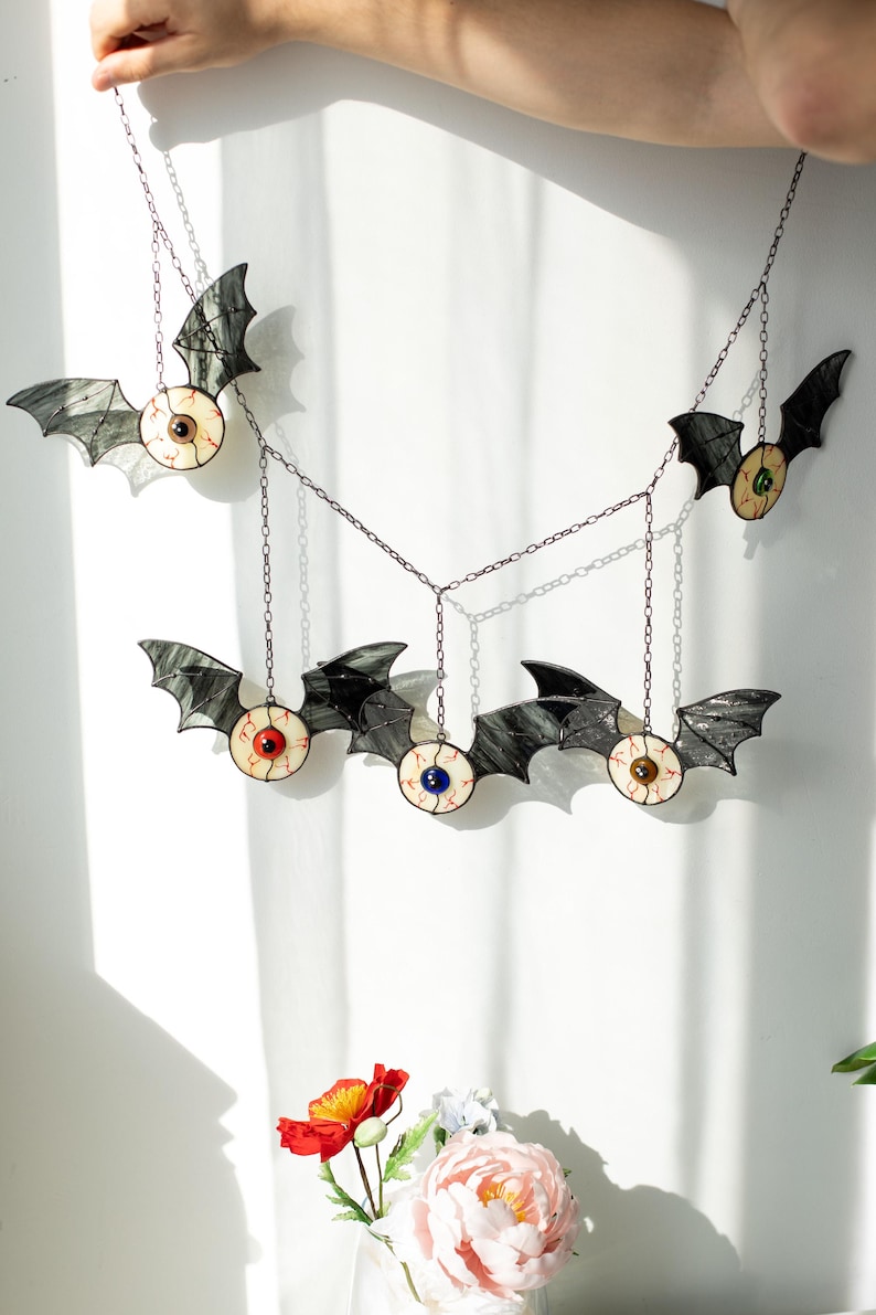 May include: A hanging mobile featuring five stained glass bats, each with a central eyeball design. The bats have dark grey wings and eyes with red, blue, green, brown, and black irises, all connected by thin metal chains for suspension.