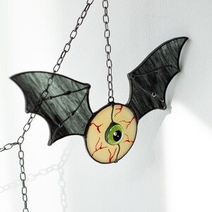 May include: A stained glass hanging ornament shaped like a bat with dark grey wings and a central circular eye. The eye features a pale yellow sclera with red veins, a bright green iris, and a black pupil. It is suspended by a silver metal chain.