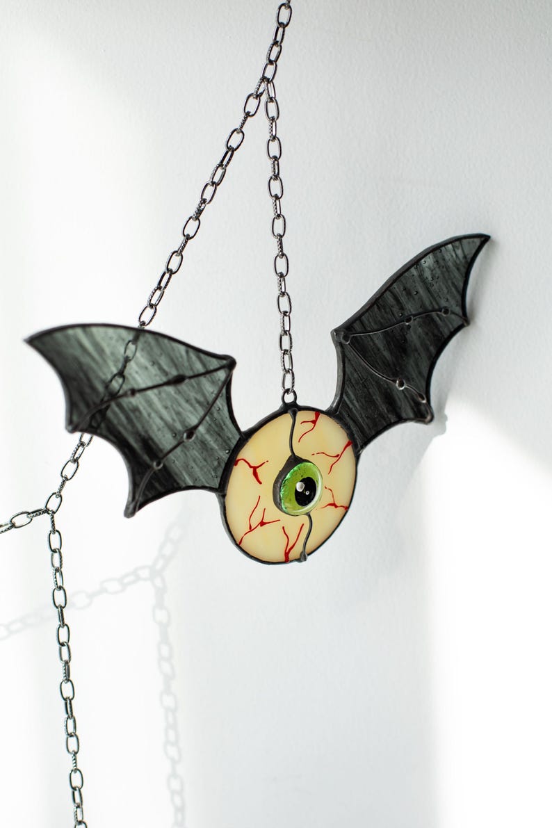 May include: A stained glass hanging ornament shaped like a bat with dark grey wings and a central circular eye. The eye features a pale yellow sclera with red veins, a bright green iris, and a black pupil. It is suspended by a silver metal chain.