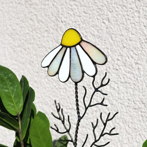 K&ouml;nnte beinhalten: Eine Glasmalerei-G&auml;nsebl&uuml;mchen-Blume mit gelber Mitte und wei&szlig;en Bl&uuml;tenbl&auml;ttern auf einem schwarzen Metallstiel mit Verzweigungen. Die Blume befindet sich vor einer gr&uuml;nen Pflanze.