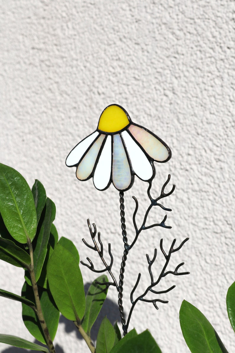 K&ouml;nnte beinhalten: Eine Glasmalerei-G&auml;nsebl&uuml;mchen-Blume mit gelber Mitte und wei&szlig;en Bl&uuml;tenbl&auml;ttern auf einem schwarzen Metallstiel mit Verzweigungen. Die Blume befindet sich vor einer gr&uuml;nen Pflanze.