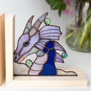 May include: Stained glass dragon head bookends. The dragon features shades of purple, blue, and green with a wooden base. The dragon's head is detailed with a whimsical design, and the bookends are a decorative and functional item.