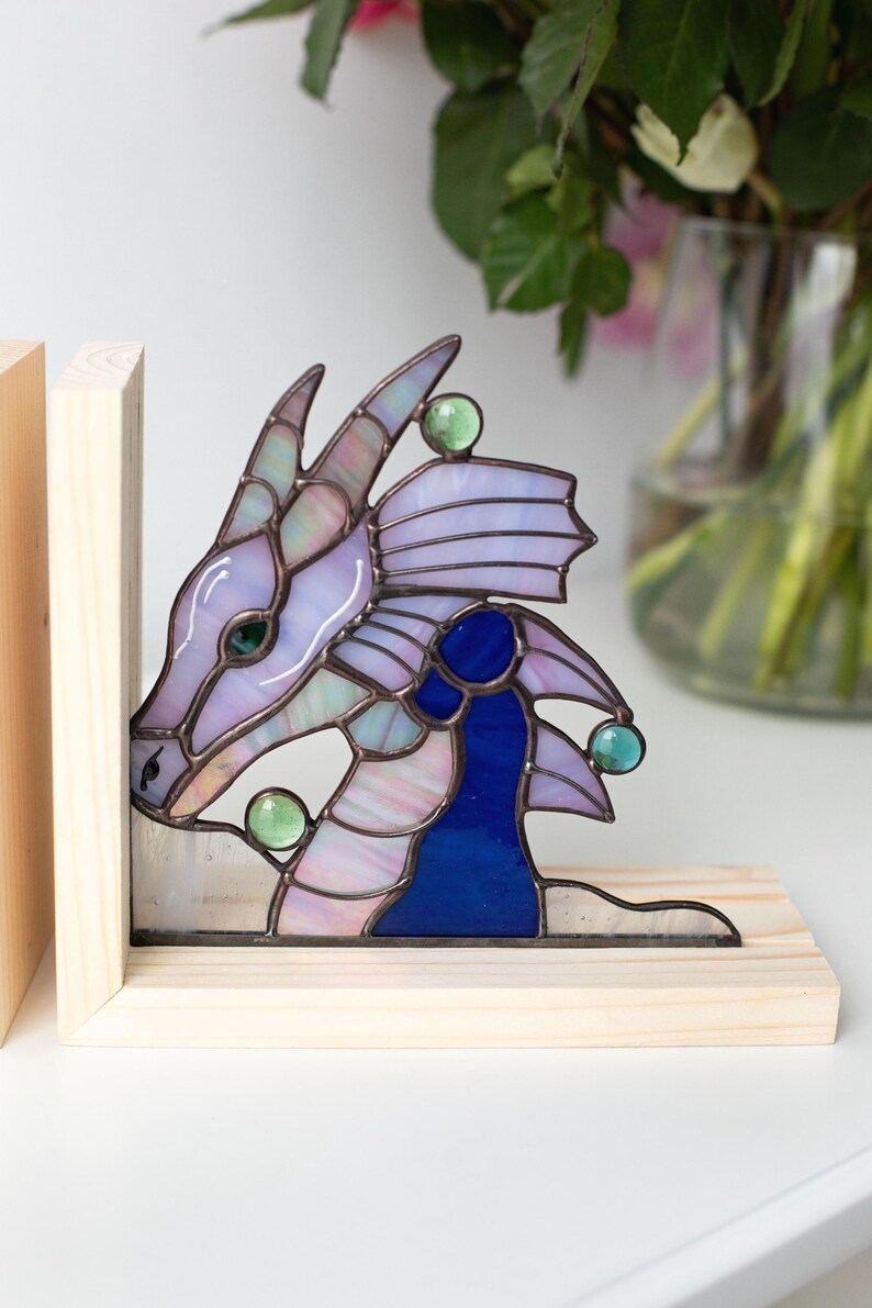 May include: Stained glass dragon head bookends. The dragon features shades of purple, blue, and green with a wooden base. The dragon's head is detailed with a whimsical design, and the bookends are a decorative and functional item.