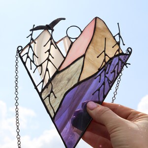May include: A stained glass window hanging with a chain. The window depicts a mountain range with a purple, blue, green, and pink color scheme. There are trees on either side of the mountain range.