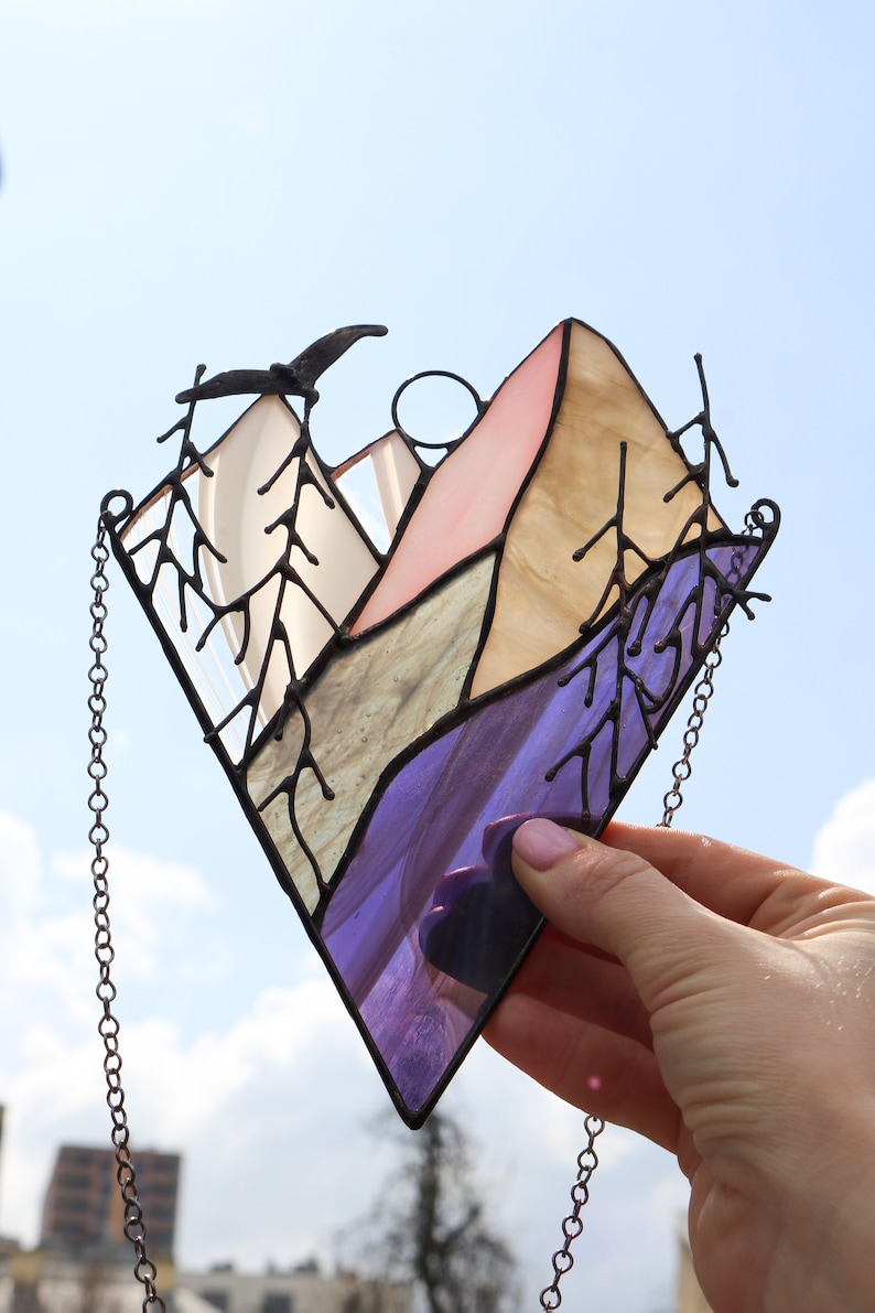 May include: A stained glass window hanging with a chain. The window depicts a mountain range with a purple, blue, green, and pink color scheme. There are trees on either side of the mountain range.