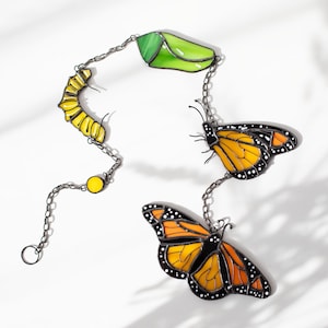 May include: A decorative chain featuring a caterpillar, chrysalis, and two monarch butterflies. The chain links are silver-colored. The caterpillar is yellow, the chrysalis is green, and the butterflies have orange and black wings.