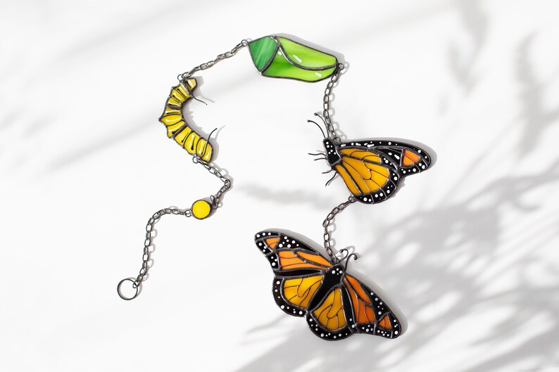 May include: A decorative chain featuring a caterpillar, chrysalis, and two monarch butterflies. The chain links are silver-colored. The caterpillar is yellow, the chrysalis is green, and the butterflies have orange and black wings.