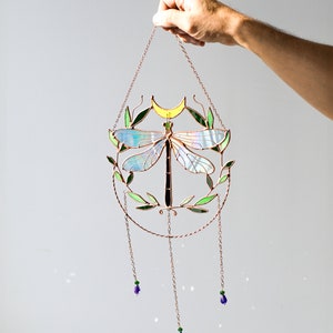 May include: A stained glass dragonfly suncatcher with a crescent moon and green leaves. The suncatcher is hanging from a copper chain with three purple glass beads.
