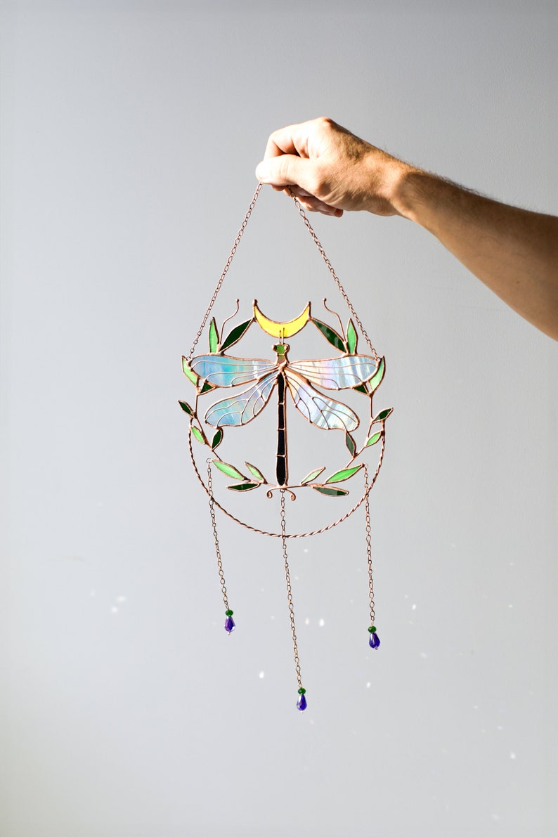 May include: A stained glass dragonfly suncatcher with a crescent moon and green leaves. The suncatcher is hanging from a copper chain with three purple glass beads.