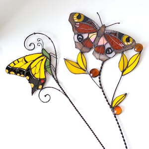 Può includere: Due picchetti decorativi da giardino a forma di farfalla. Una farfalla è gialla e nera, l'altra è marrone, rossa e gialla. Entrambe hanno foglie gialle e perline arancioni. I picchetti sono in metallo.