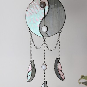 May include: A stained glass yin yang symbol with a gray and iridescent blue and pink design hangs from a chain with three iridescent blue and pink feathers and white beads.