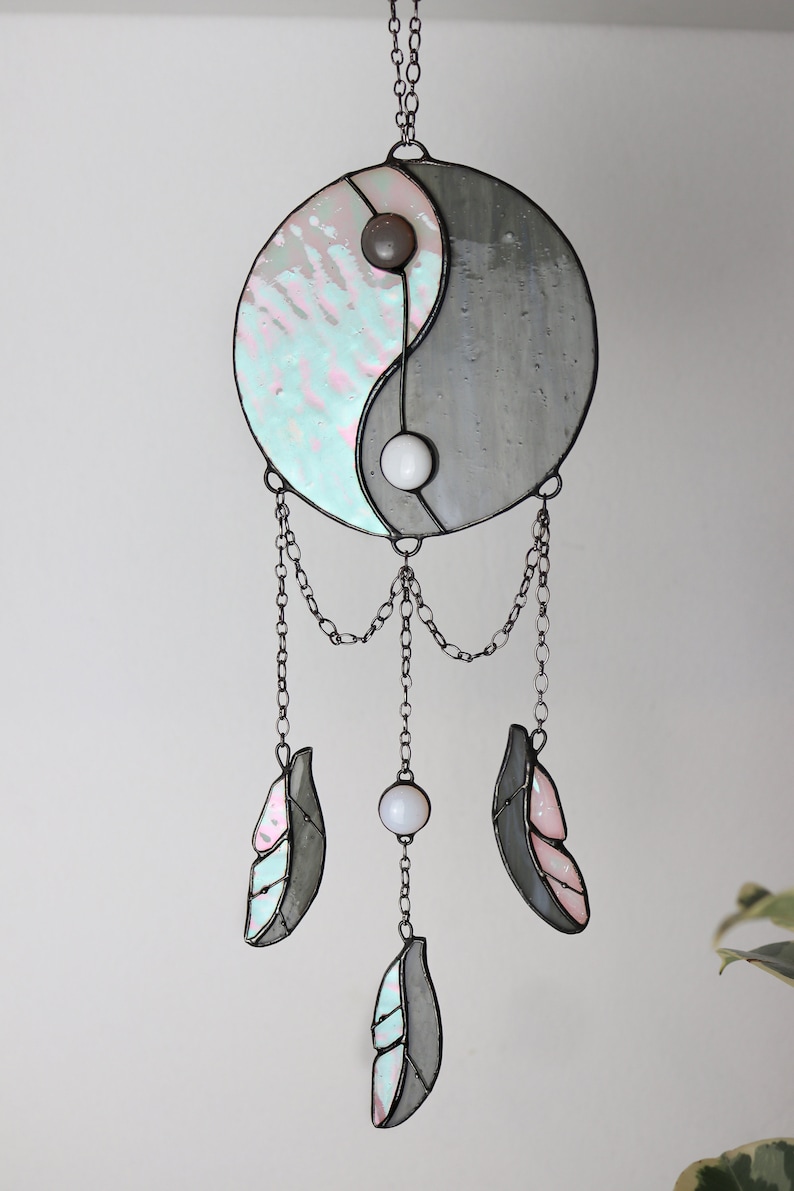 May include: A stained glass yin yang symbol with a gray and iridescent blue and pink design hangs from a chain with three iridescent blue and pink feathers and white beads.