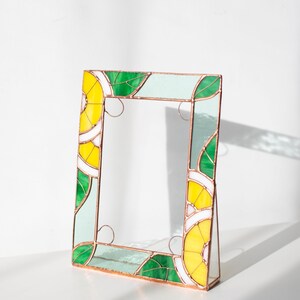 May include: A stained glass picture frame with a vibrant design. The frame features yellow, green, white, and light blue glass pieces, arranged in a geometric pattern. The frame is supported by a small stand, and the edges are outlined with copper.