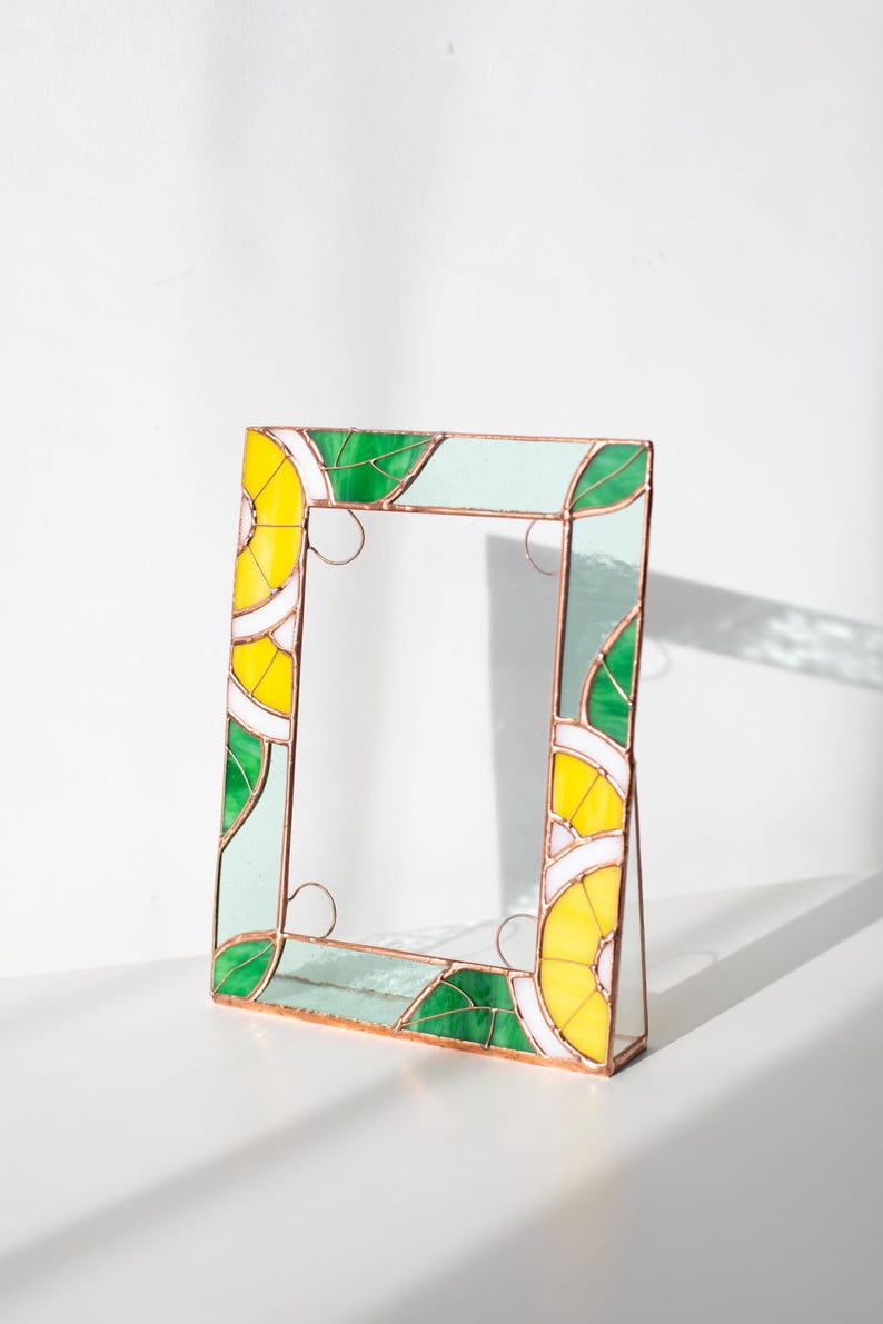 May include: A stained glass picture frame with a vibrant design. The frame features yellow, green, white, and light blue glass pieces, arranged in a geometric pattern. The frame is supported by a small stand, and the edges are outlined with copper.