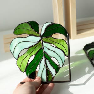 K&ouml;nnte beinhalten: Monstera-Blatt aus Buntglas in Gr&uuml;n- und Wei&szlig;t&ouml;nen, mit einem dunklen Metallrahmen. Das Blattdesign weist verschiedene Gr&uuml;nt&ouml;ne und wei&szlig;es Glas auf und schafft ein lebendiges und dekoratives St&uuml;ck. Ein einzigartiger Einrichtungsgegenstand.