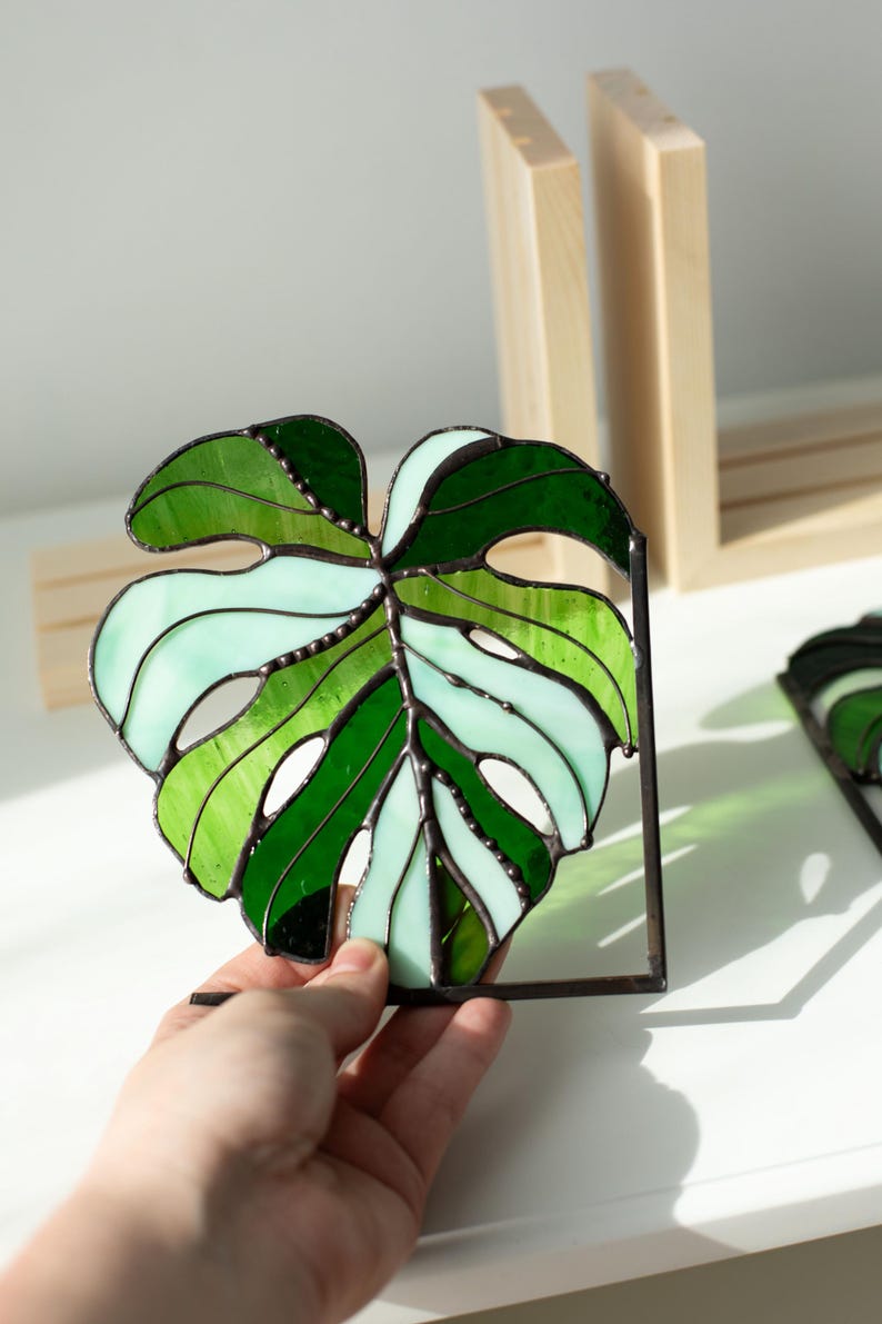 K&ouml;nnte beinhalten: Monstera-Blatt aus Buntglas in Gr&uuml;n- und Wei&szlig;t&ouml;nen, mit einem dunklen Metallrahmen. Das Blattdesign weist verschiedene Gr&uuml;nt&ouml;ne und wei&szlig;es Glas auf und schafft ein lebendiges und dekoratives St&uuml;ck. Ein einzigartiger Einrichtungsgegenstand.