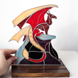 Op de afbeelding: Gebrandschilderde drakenbeeld in rood, oranje en gele tinten, bovenop een zwarte en paarse rotsformatie. De draak staat op een donkere houten basis, wat een opvallend contrast cre&euml;ert. De vleugels zijn gespreid, de draak zit.